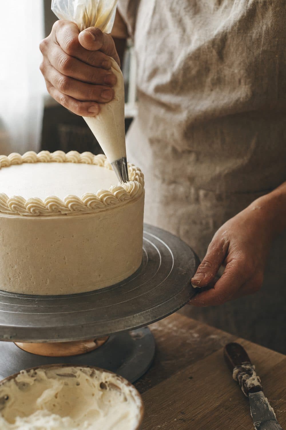 A pastry chef piping a buttercream border onto an elegant single-tier cake by hand